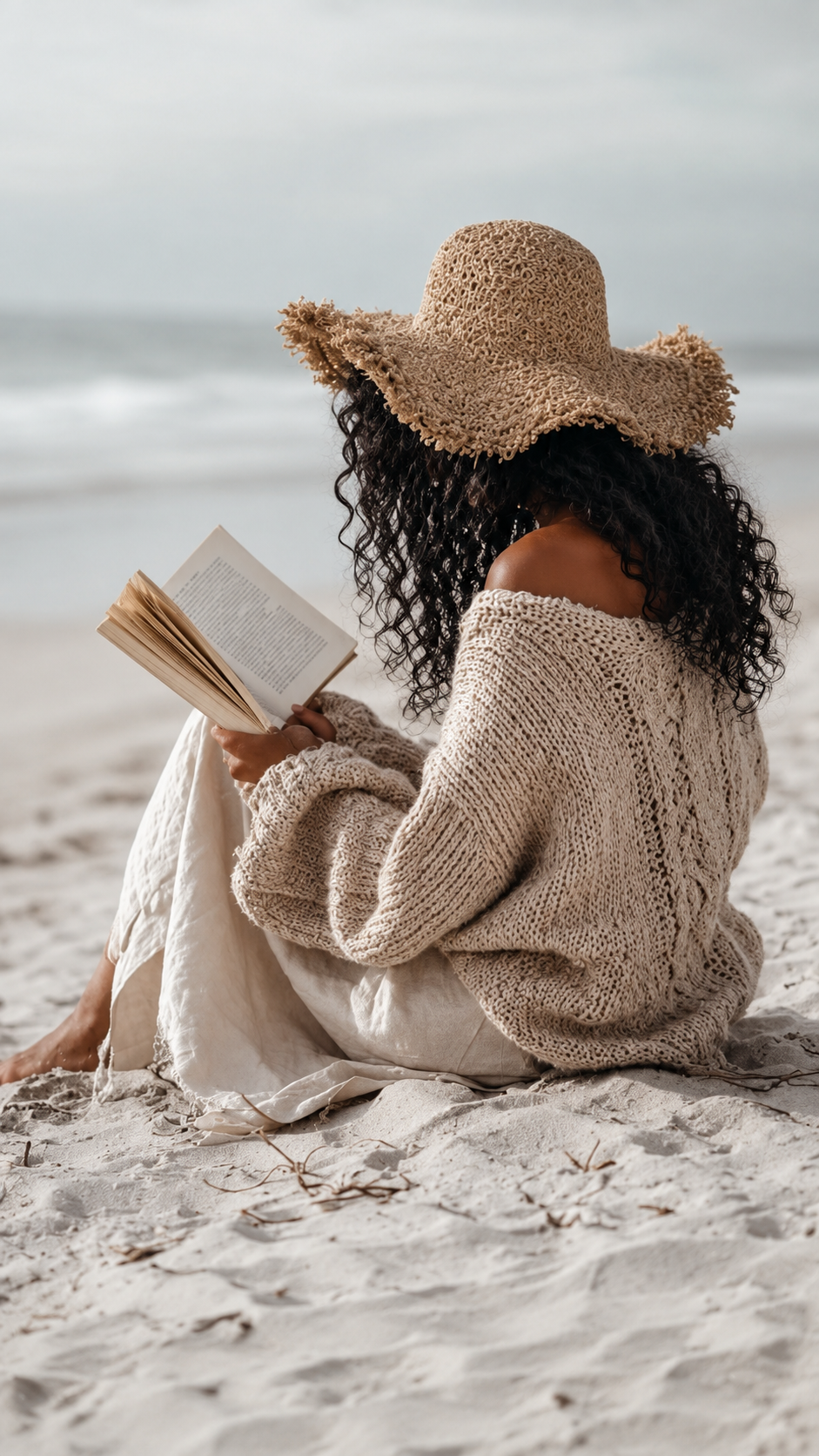 A woman seated alone on an empty beach, back to the camera, reading